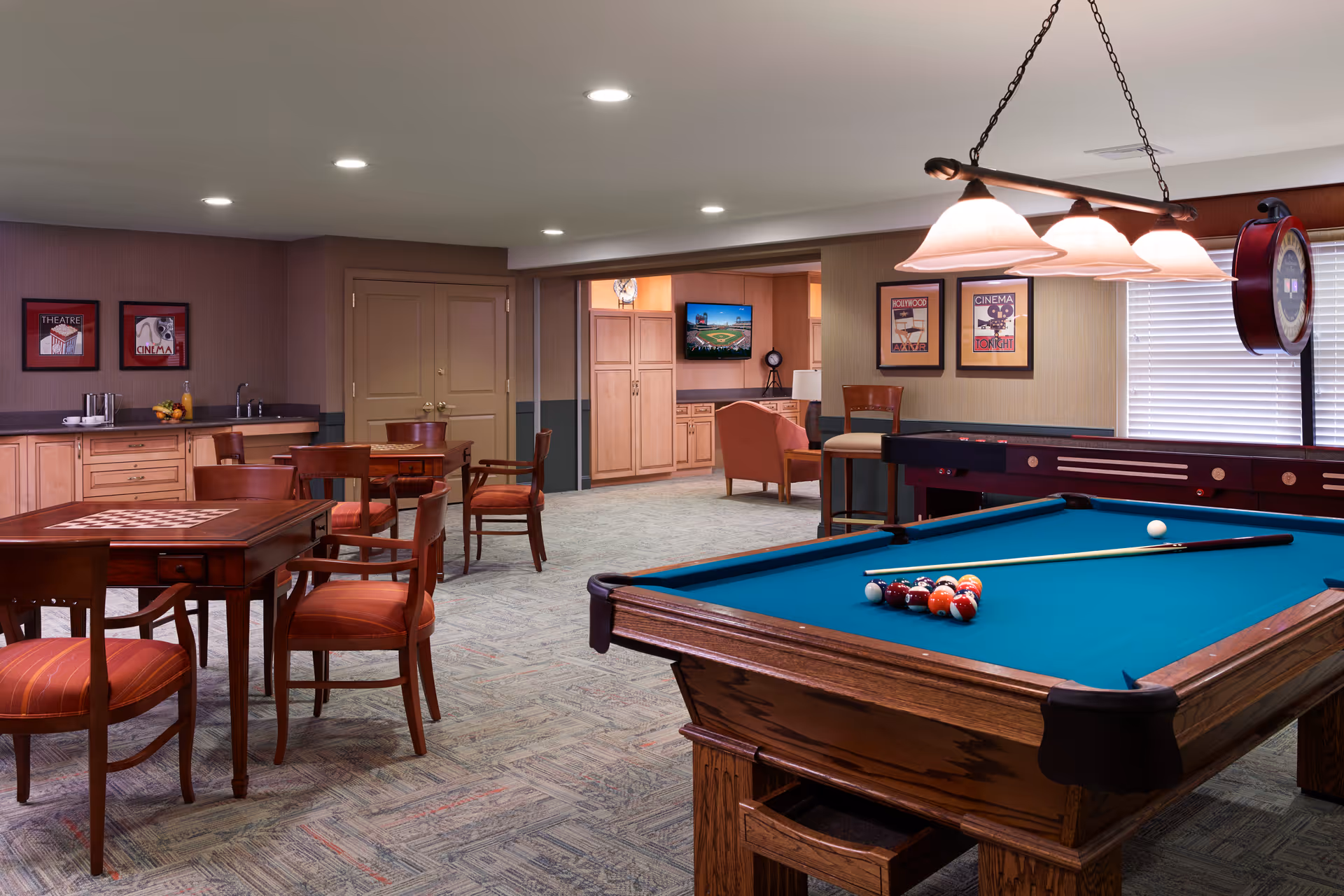 A recreational room with a pool table in the foreground, a shuffleboard table, and several wooden tables and chairs with red cushions. The room has beige walls with framed movie-themed artwork, a small kitchenette area with cabinets and a sink, and a television mounted on the far wall. The lighting is provided by recessed ceiling lights and a hanging lamp above the pool table.