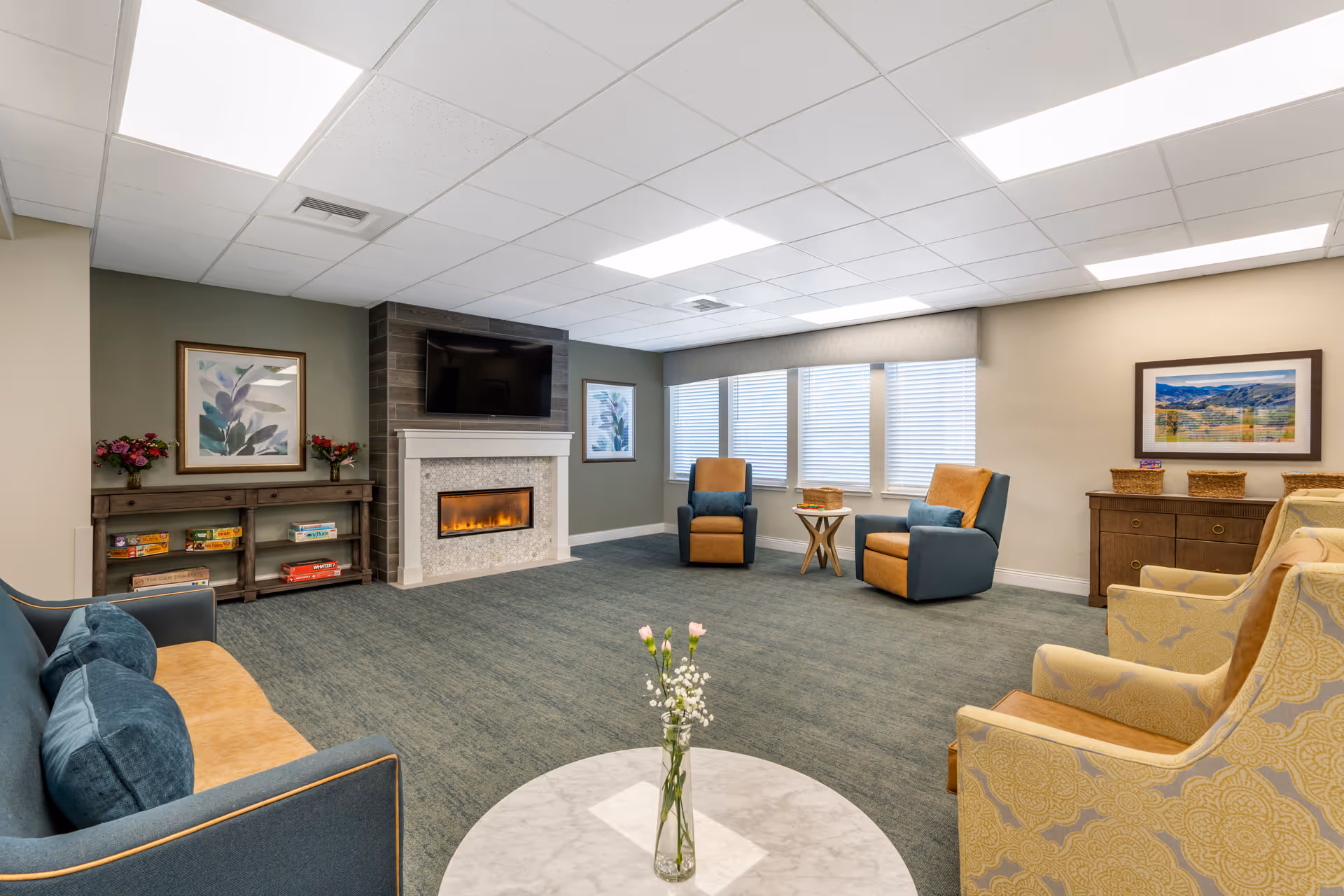 A spacious and well-lit living room with a blue and tan sofa, two blue and tan armchairs, and two yellow patterned armchairs arranged around a round marble coffee table with a vase of flowers. The room features a modern electric fireplace with a TV mounted above it, large windows with blinds, framed artwork on the walls, and shelves with board games and decorative baskets.