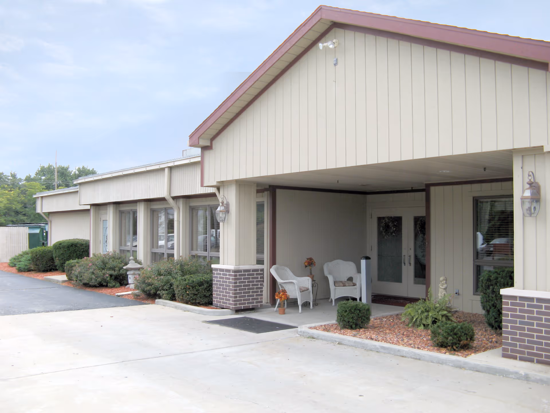 Covered front entrance of a single-story senior living building with chairs, planters, and landscaping.