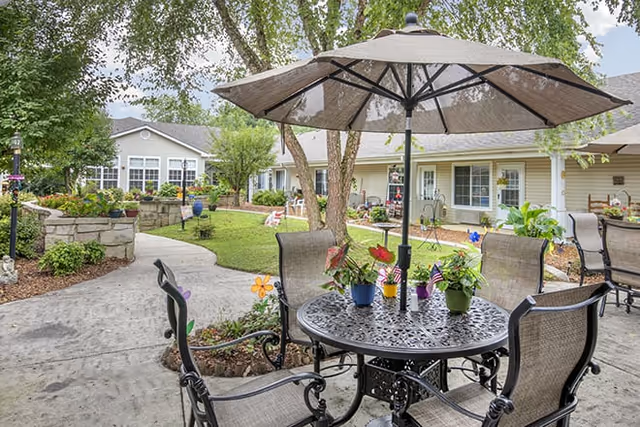 Outdoor patio area at Brookdale Tullahoma featuring a round metal table with four chairs and a large umbrella. The patio is surrounded by a garden with plants, flowers, and trees, with residential-style buildings in the background.