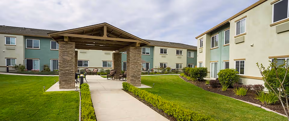 Outdoor view of Eskaton Granite Bay senior living facility showing a covered walkway with stone pillars, surrounded by green lawns and shrubs, with two-story residential buildings in the background under a partly cloudy sky.
