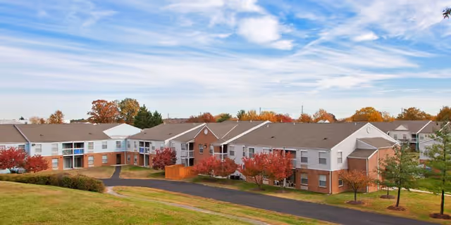 Exterior view of a two-story brick-and-siding senior living complex surrounded by lawns and autumn trees under a blue sky.