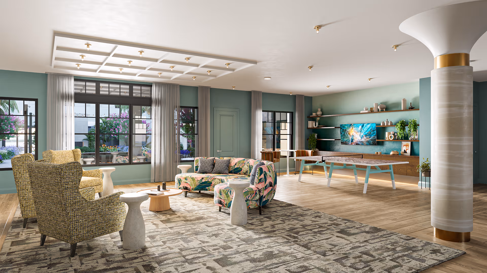 A spacious and modern living room area with large windows showing an outdoor garden. The room features a patterned area rug, two yellow armchairs, a floral-patterned sofa, small white side tables, a round wooden coffee table, and a ping pong table. The walls are painted teal with shelves holding decorative items and a mounted flat-screen TV. A large column with gold accents is visible on the right side.