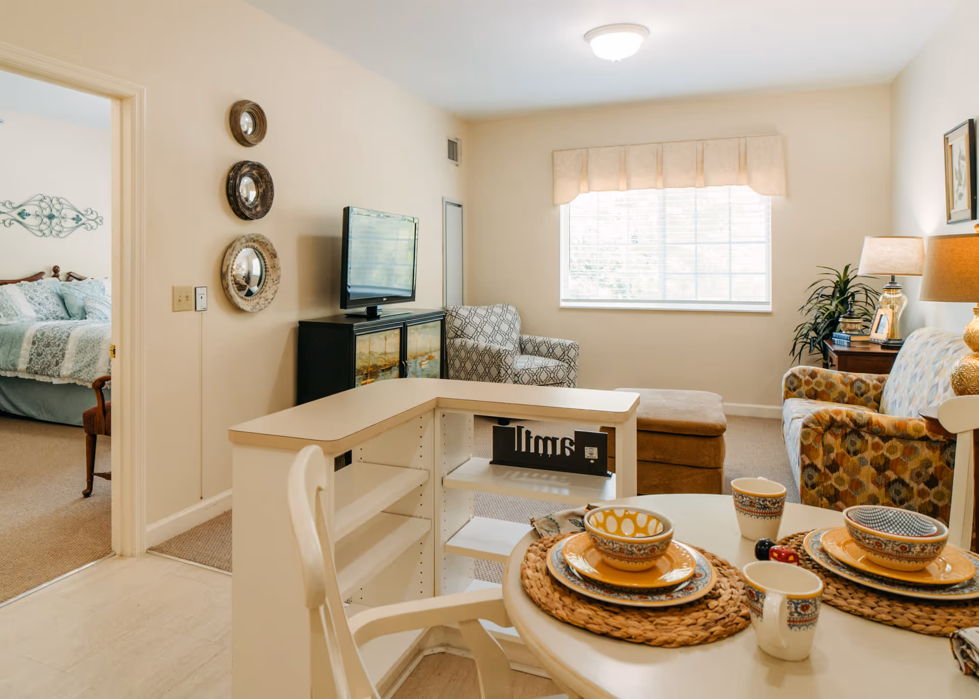 Bright living area with a dining table set for two, seating, a TV console, and an adjacent bedroom visible through an open door.