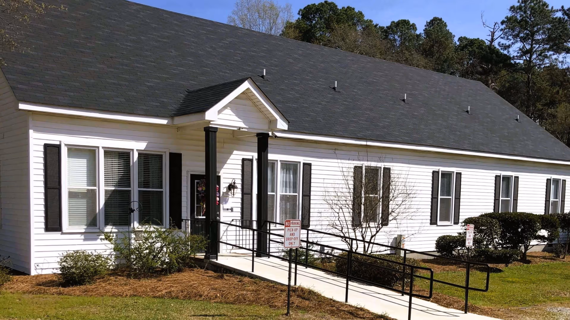 Exterior view of a single-story white building with black shutters and a dark shingled roof. The entrance features a small covered porch with two black columns and a wheelchair ramp leading up to the door. There are bushes and small trees planted around the building, and a grassy lawn in front. A sign near the ramp reads 'No Parking Pick-Up and Drop-Off Only.'