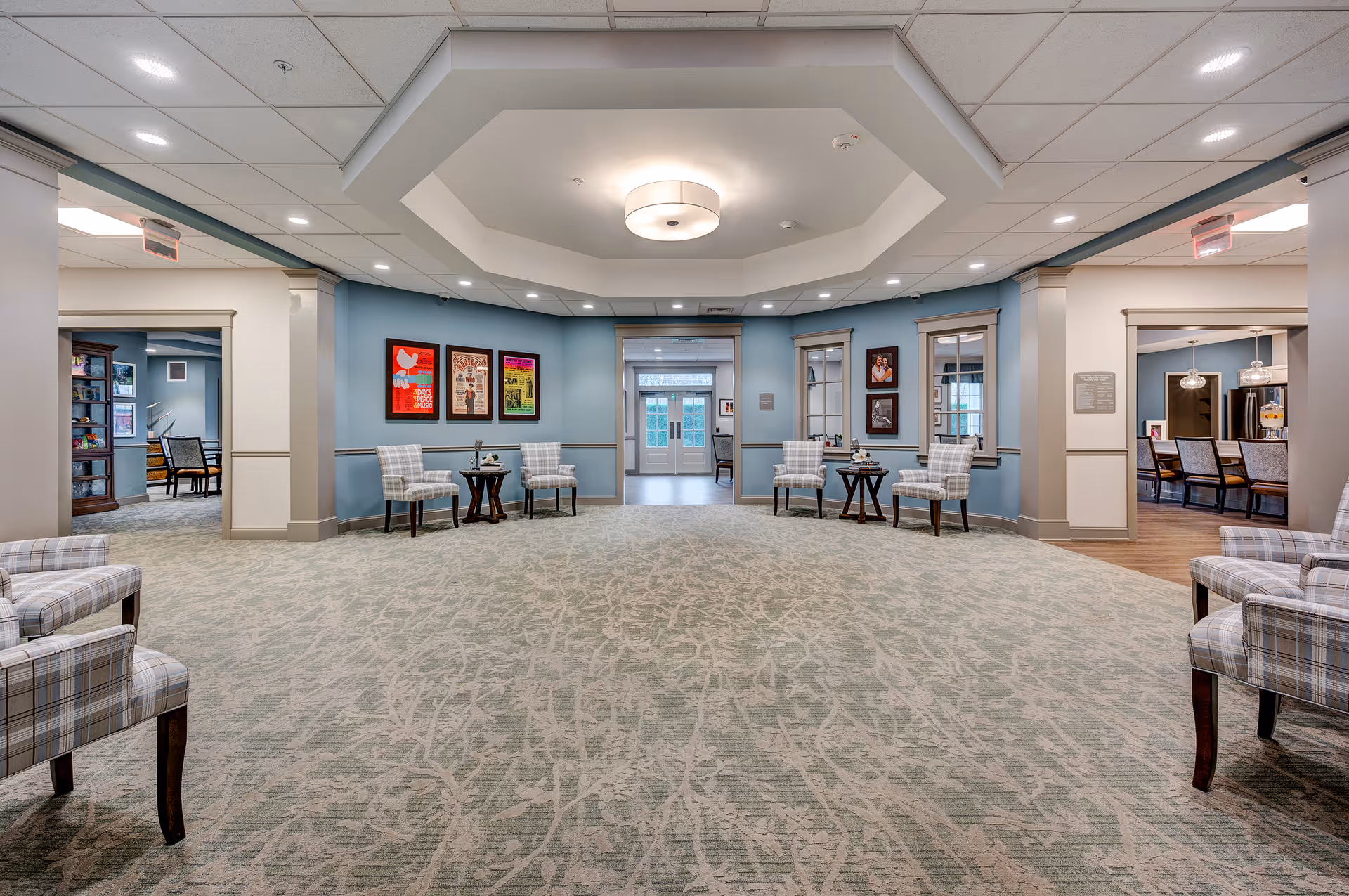 Spacious and well-lit common area in a senior living facility with light blue walls, patterned carpet, and several plaid upholstered chairs arranged in pairs with small tables. The ceiling features a modern light fixture and recessed lighting. Framed artwork and photographs decorate the walls. Doorways lead to other rooms, including a dining area with a long table and chairs.