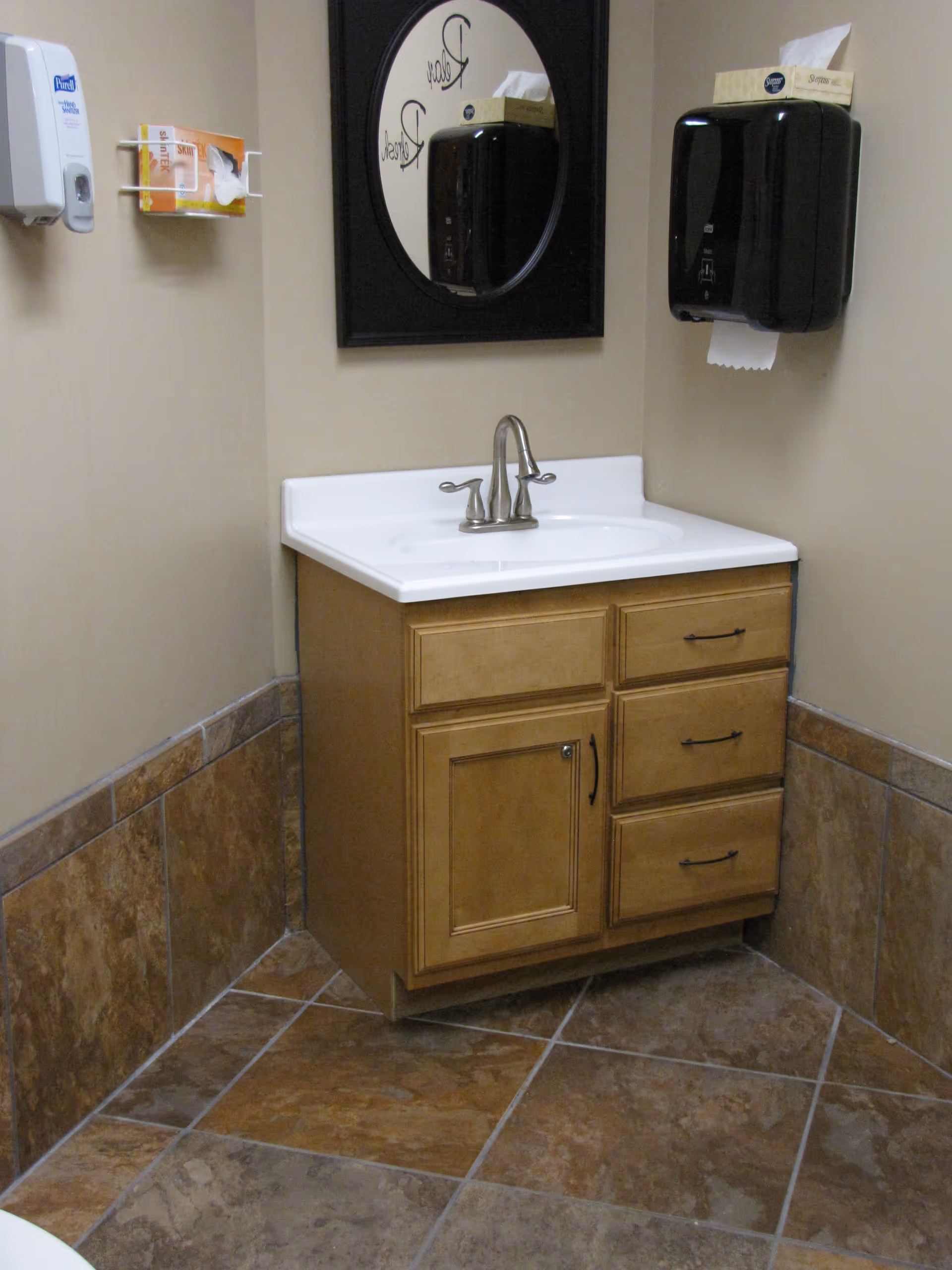 A small bathroom corner with a wooden vanity cabinet topped with a white sink and silver faucet. Above the sink is a round mirror with a black frame. On the wall to the right is a black paper towel dispenser with a tissue box on top, and on the left wall is a white soap dispenser and a box of tissues mounted on the wall. The floor and lower walls are tiled in brown and beige tones.