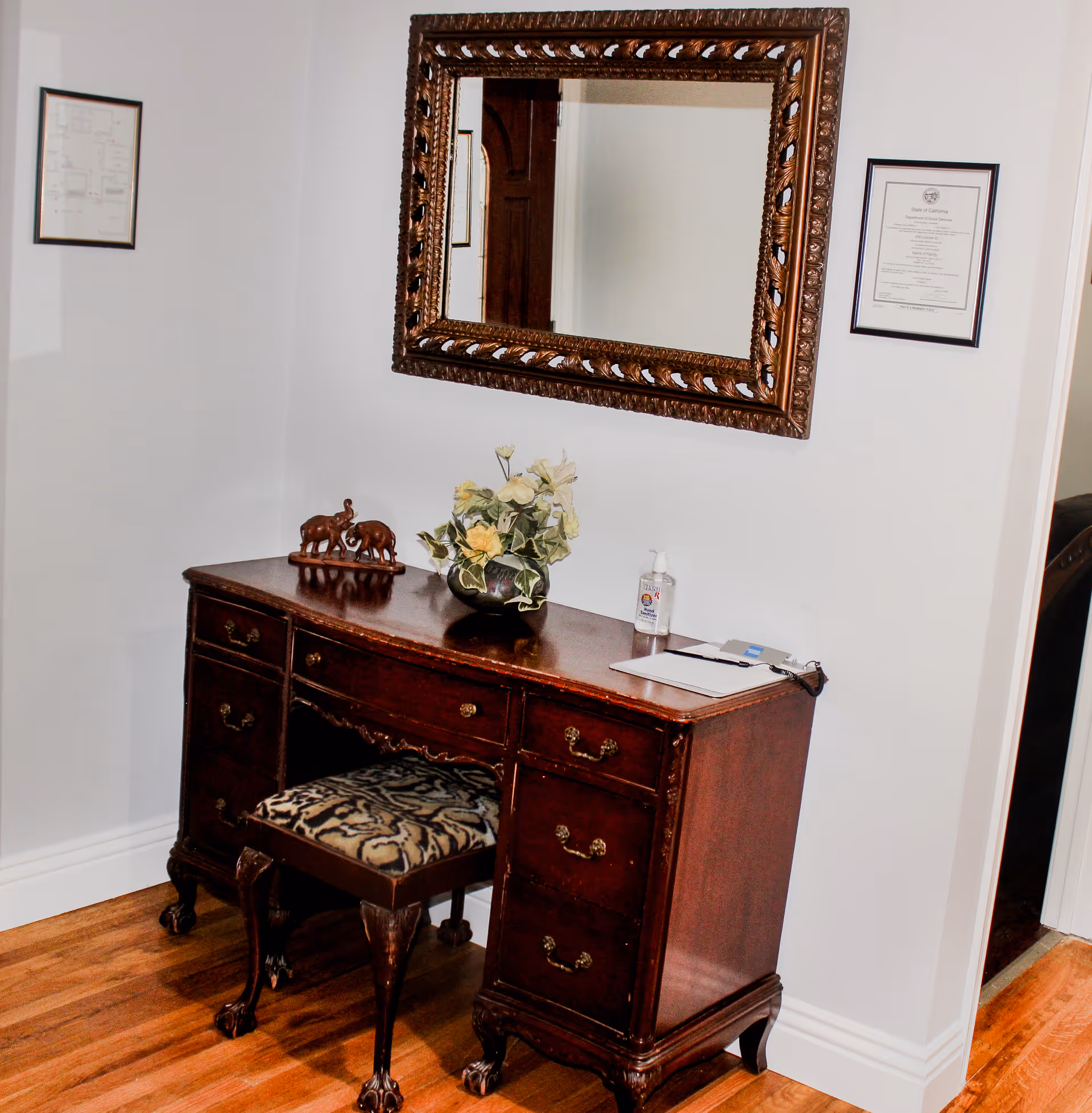 A wooden vanity desk with multiple drawers and an upholstered stool with a patterned fabric seat. On top of the desk are a decorative elephant figurine, a vase with yellow and white flowers, a bottle of hand sanitizer, and some papers. Above the desk is a large ornate framed mirror. The walls are white with two framed documents hanging on either side of the mirror. The floor is wooden.