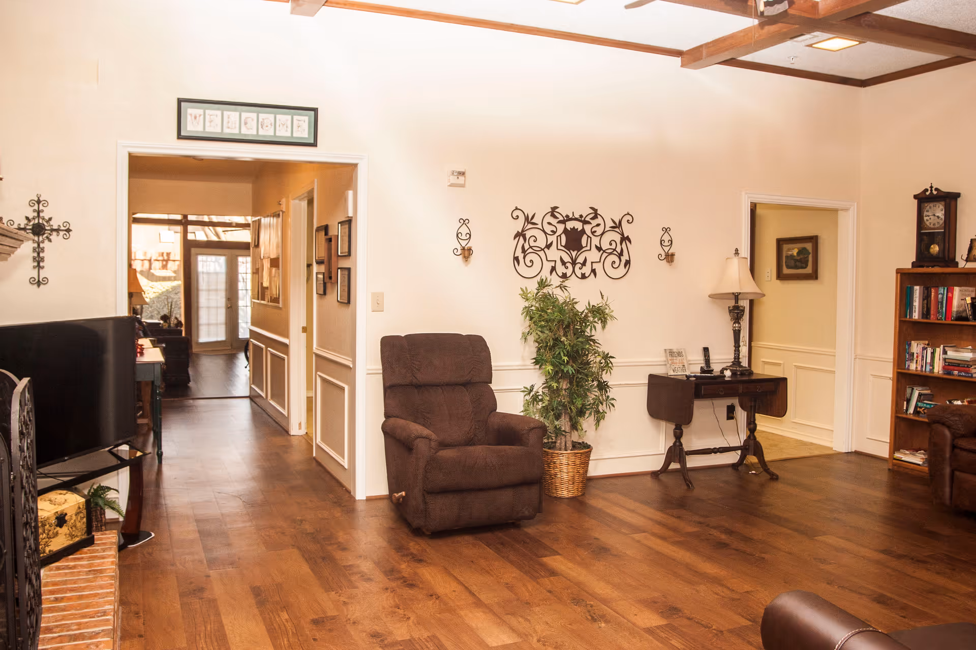 A cozy living room area with wooden flooring, a brown recliner chair, a small table with a lamp, a bookshelf filled with books, and decorative wall art. There is a hallway leading to a door with glass panels, and the room has a warm, inviting atmosphere with beige walls and wooden ceiling beams.