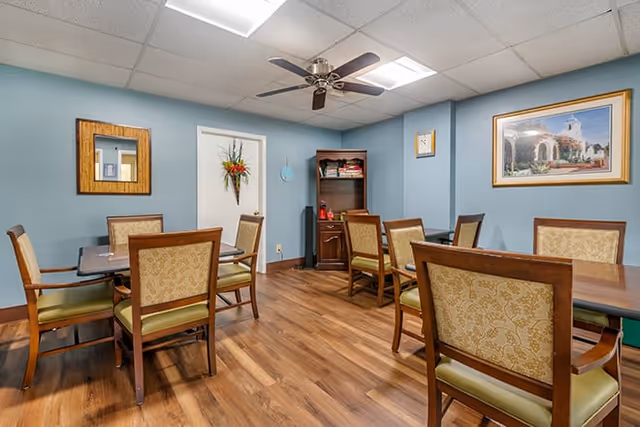 A small dining room with several wooden tables and upholstered chairs, blue walls, ceiling fan, artwork, and wood-look flooring.