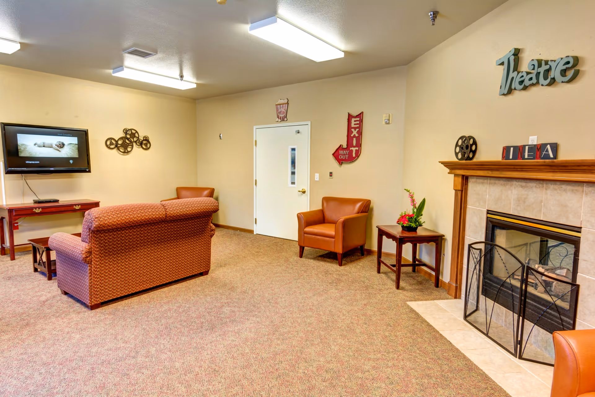 A cozy common room with a patterned sofa and two leather armchairs facing a wall-mounted TV. The room features a fireplace with a decorative screen, a small table with a flower arrangement, and wall decorations including a 'Theatre' sign and a red exit sign. The carpet is beige, and the walls are light-colored.