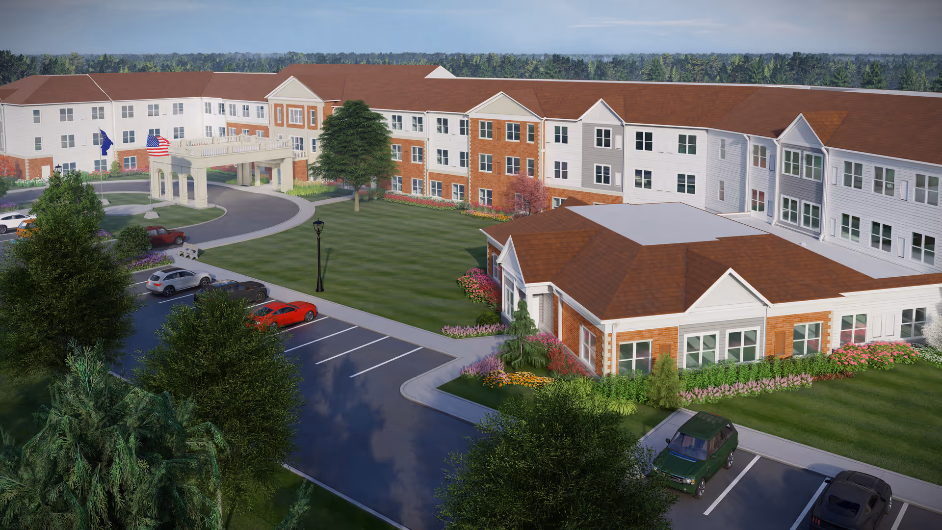 Exterior view of a large senior living facility with a three-story building featuring a mix of brick and white siding. The building surrounds a green lawn with landscaped flower beds and trees. There is a circular driveway with a covered entrance, several parked cars, and flags near the entrance.