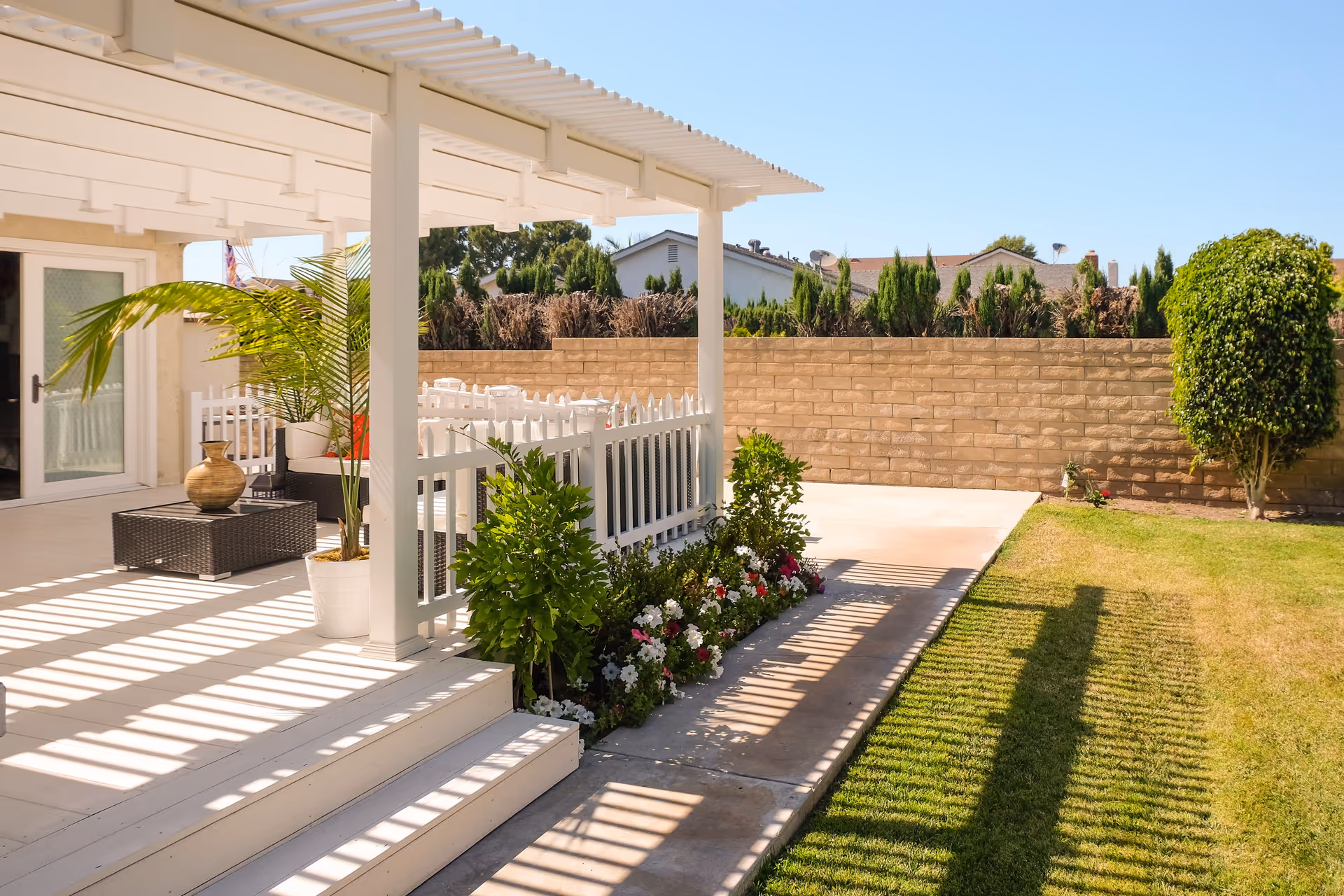 Sunlit backyard patio with a white pergola-covered deck, potted plants, a flower bed, lawn and a brick privacy wall.