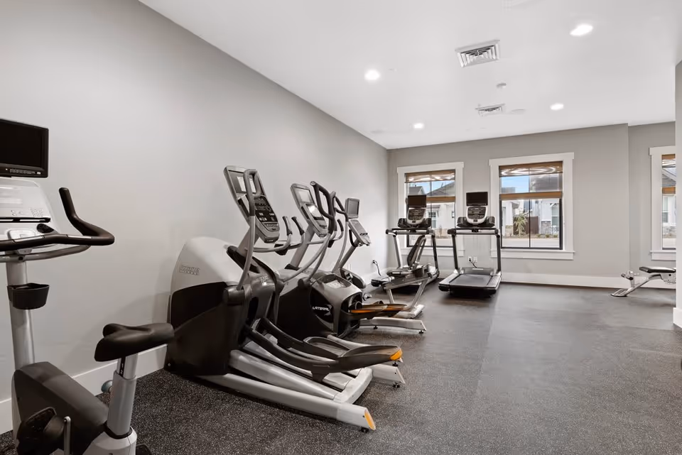 A fitness room with exercise equipment including stationary bikes, elliptical machines, and treadmills. The room has light gray walls, three windows letting in natural light, and a speckled rubber floor.