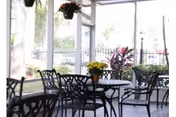 A covered outdoor patio area with black metal tables and chairs. There are potted plants on the tables and hanging plants above. The patio is enclosed with glass or clear panels, and a fence and greenery are visible outside.