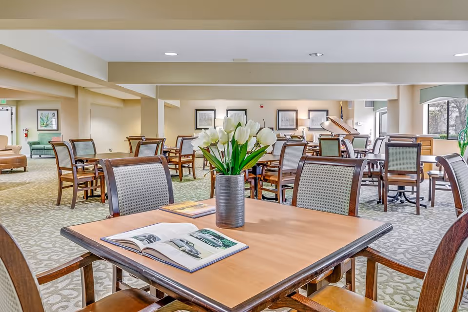 A spacious senior living common area with multiple wooden tables and chairs arranged neatly. A vase with white tulips is placed on the closest table along with an open book. The room has beige walls, patterned carpet, framed artwork on the walls, and large windows letting in natural light.