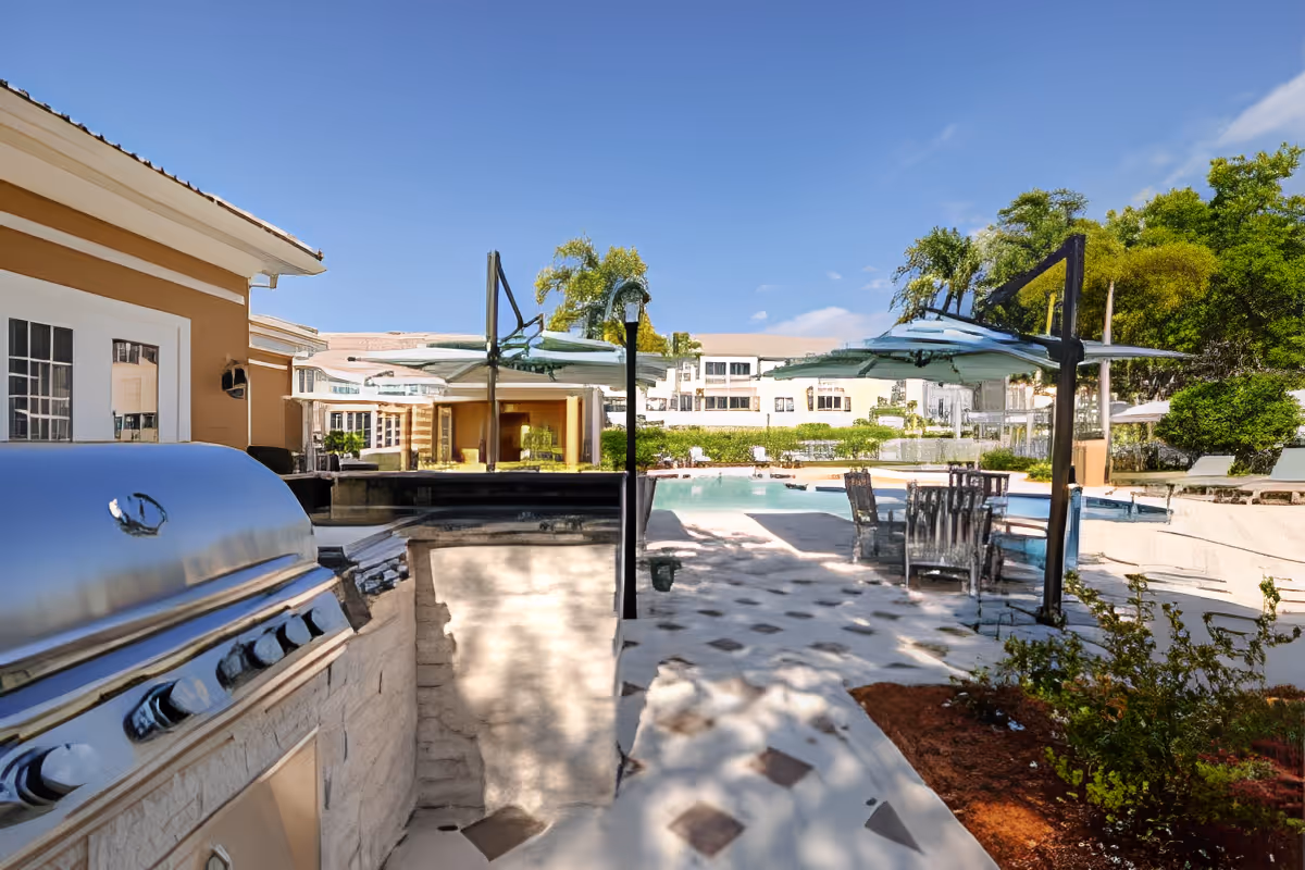 Outdoor pool and patio area with a stainless steel barbecue, umbrellas, tables and chairs in front of a residential building.