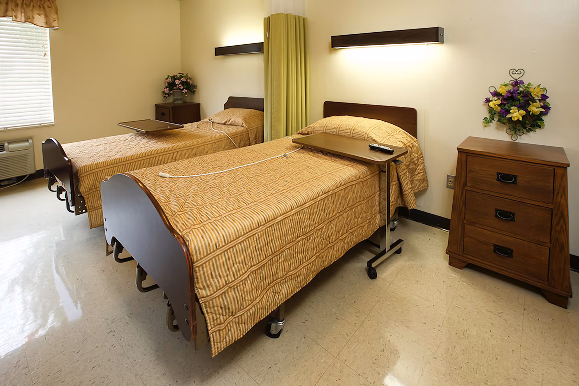 A room with two single beds covered with matching beige striped bedspreads. Each bed has a small overbed table, one with a remote control on it. There is a wooden nightstand with three drawers and a floral wall decoration above it. A window with blinds and an air conditioning unit is visible on the left side of the room.