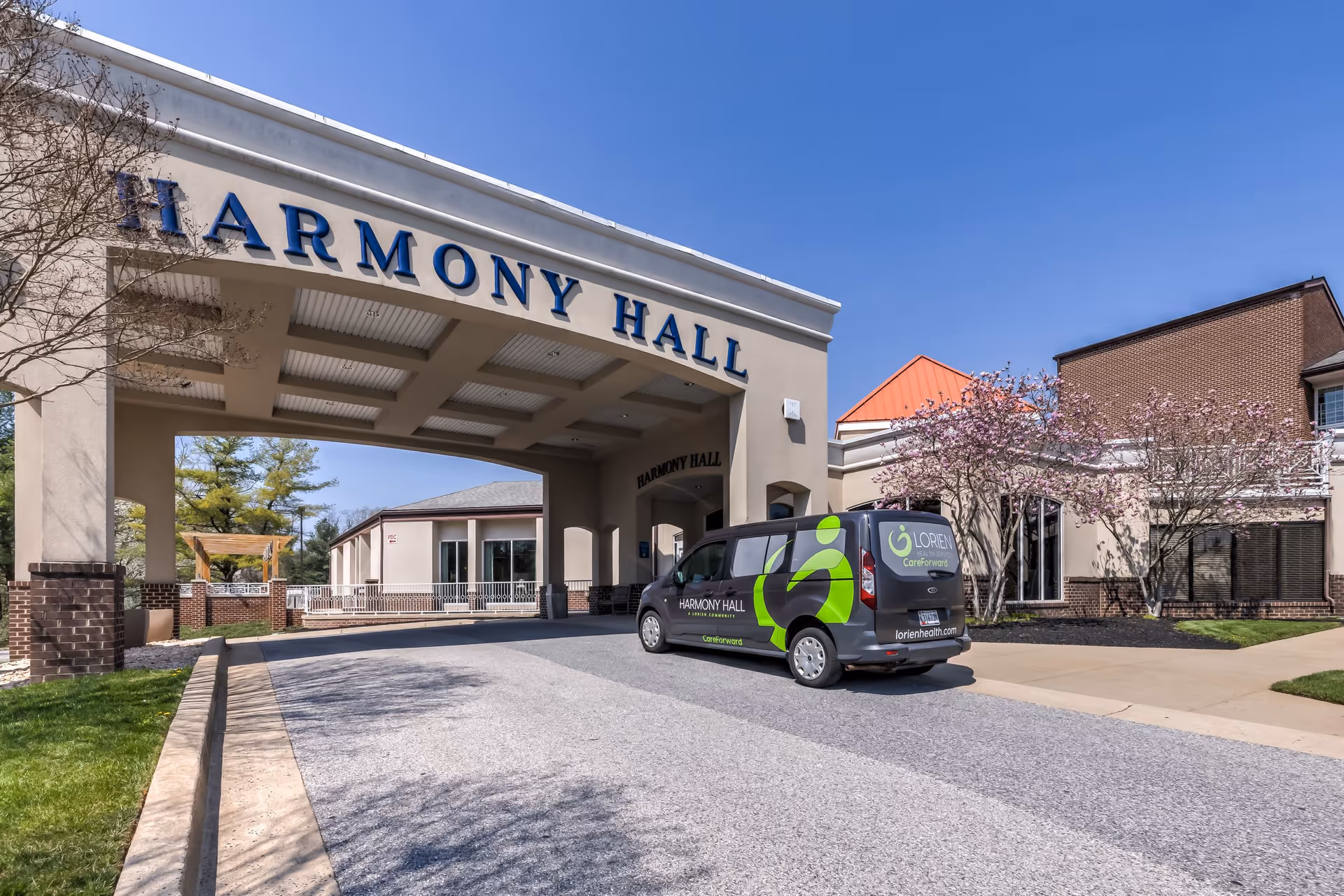 Exterior view of the entrance to Harmony Hall, a senior living facility, with a covered driveway and a Lorien Health Services van parked nearby. The building features beige walls, brick accents, and blooming trees under a clear blue sky.