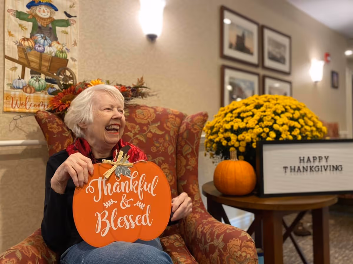 An elderly woman with white hair is sitting in a patterned armchair, smiling and holding an orange pumpkin-shaped sign that says 'Thankful & Blessed.' Next to her is a small round table with a yellow chrysanthemum plant, a small pumpkin, and a lightbox sign that reads 'HAPPY THANKSGIVING.' The background features a wall with framed pictures and a fall-themed decoration with a scarecrow and pumpkins.