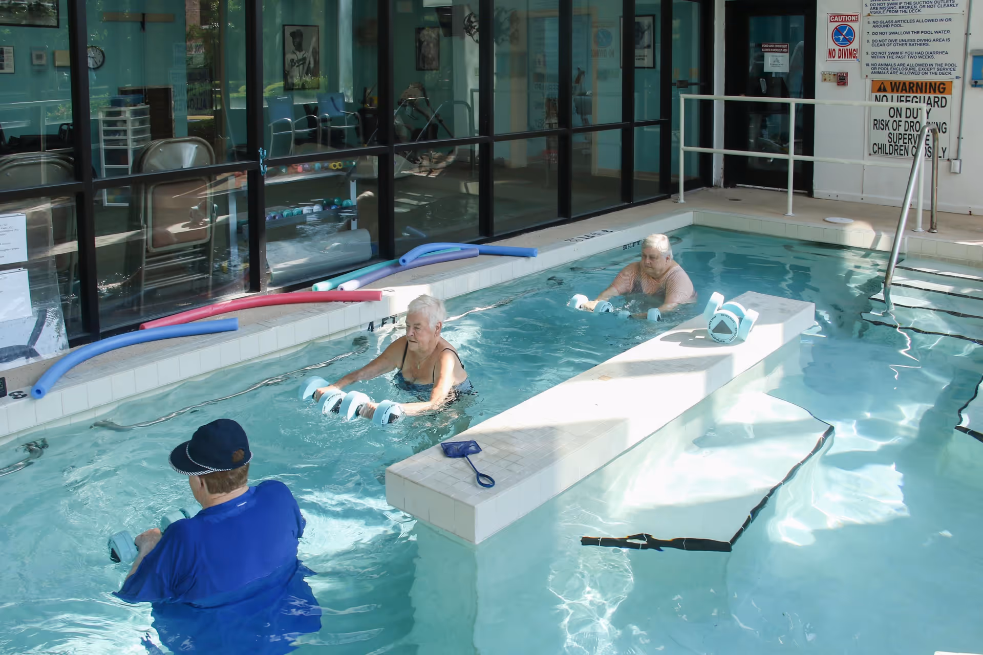 Three elderly individuals participating in water exercise in an indoor pool at a senior living facility. They are using foam dumbbells for aquatic therapy. The pool area has large windows, pool noodles on the side, and safety signs on the wall.