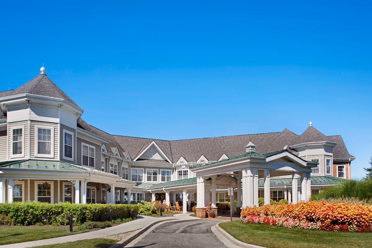 Exterior view of Sunrise of Palos Park senior living facility showing a large, two-story building with a covered entrance, manicured bushes, colorful flower beds, and a clear blue sky.