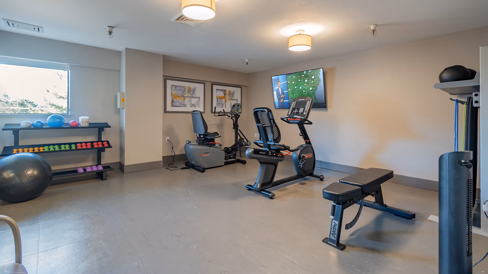A small fitness room with exercise equipment including two stationary bikes, a weight bench, a rack of colorful dumbbells, exercise balls, and resistance bands. A flat-screen TV is mounted on the wall showing a weather forecast. The room has neutral-colored walls and ceiling lights.