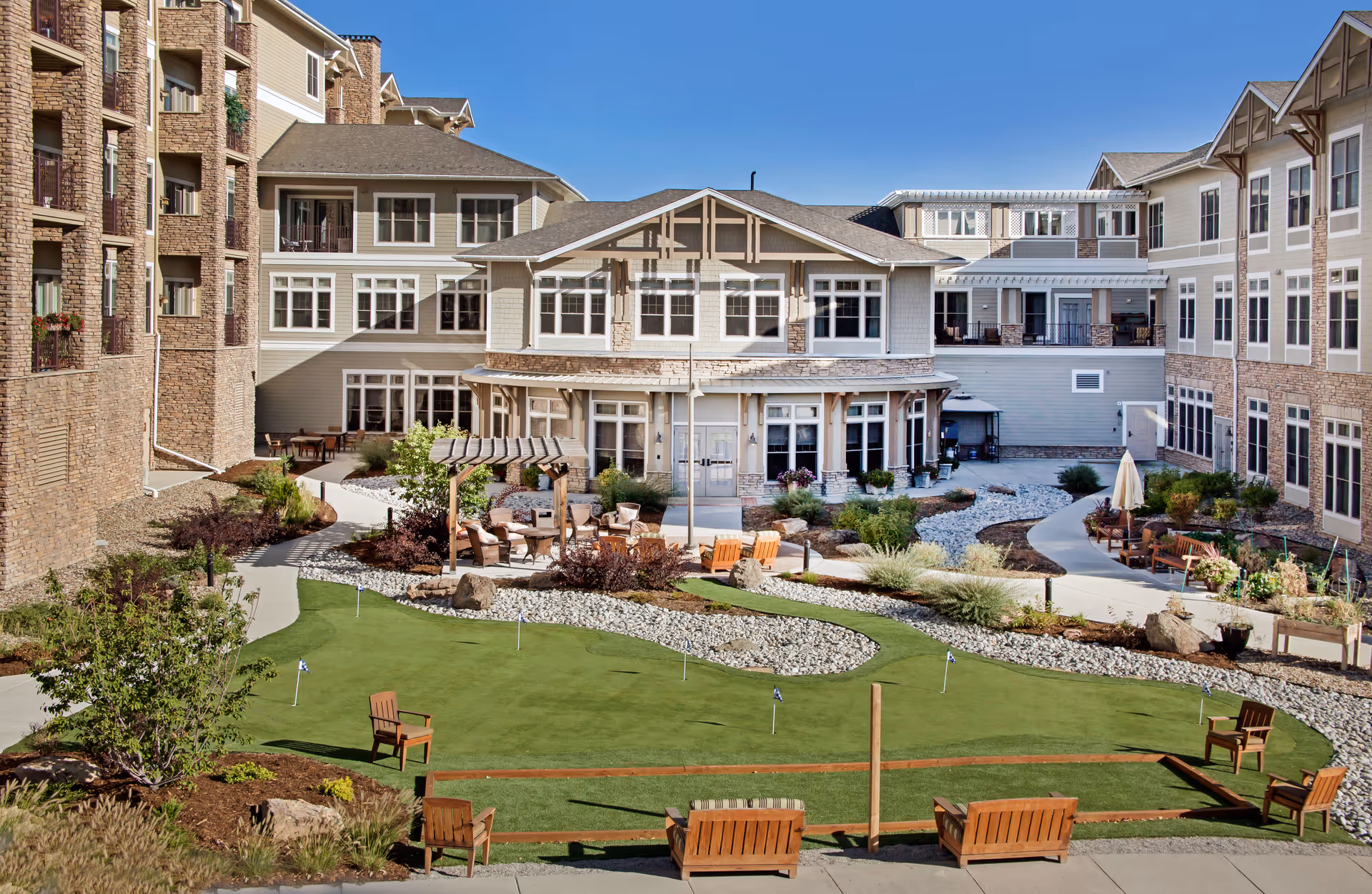 Outdoor courtyard area of a senior living facility with a putting green, wooden chairs, and a pergola with seating. The courtyard is surrounded by multi-story buildings with numerous windows and balconies. There are landscaped plants, shrubs, and pathways throughout the area under a clear blue sky.