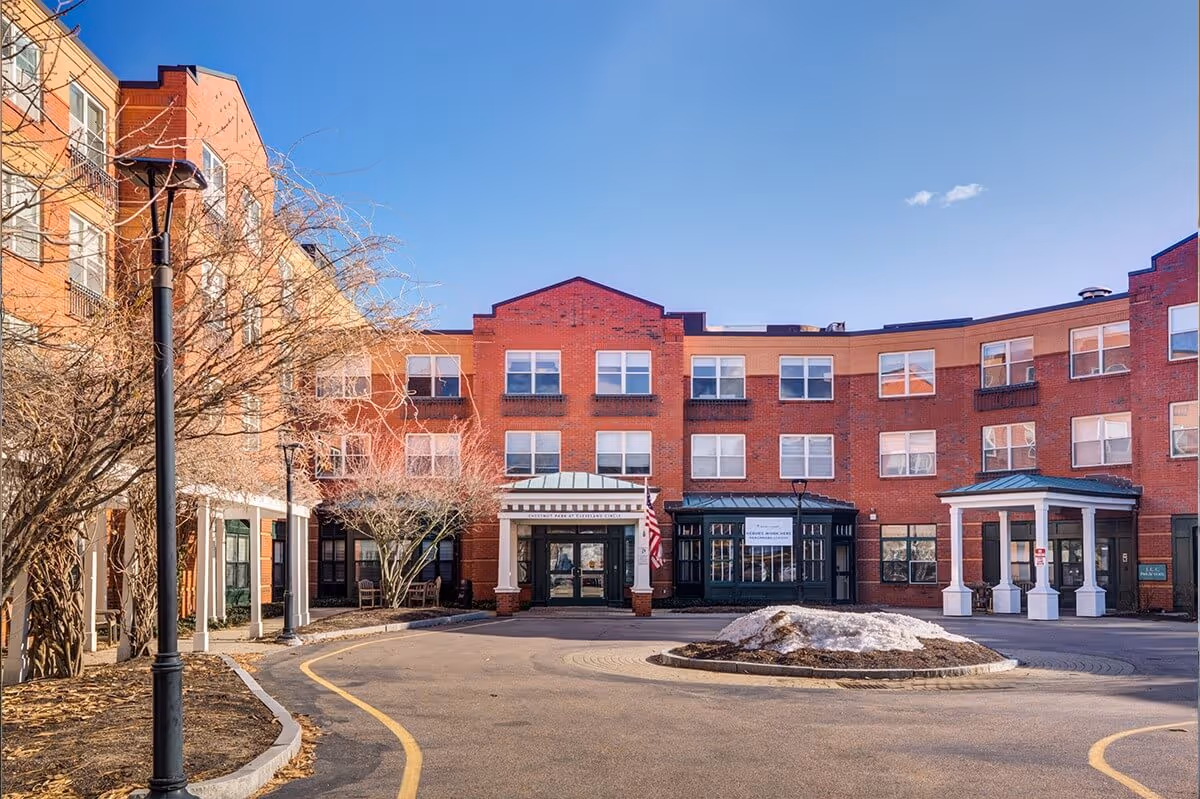 Front exterior view of a multi-story senior living facility named Chestnut Park at Cleveland Circle, featuring a red brick facade, multiple windows, a covered entrance with white columns, and a circular driveway with a small landscaped island in the center. Leafless trees and a clear blue sky are visible.