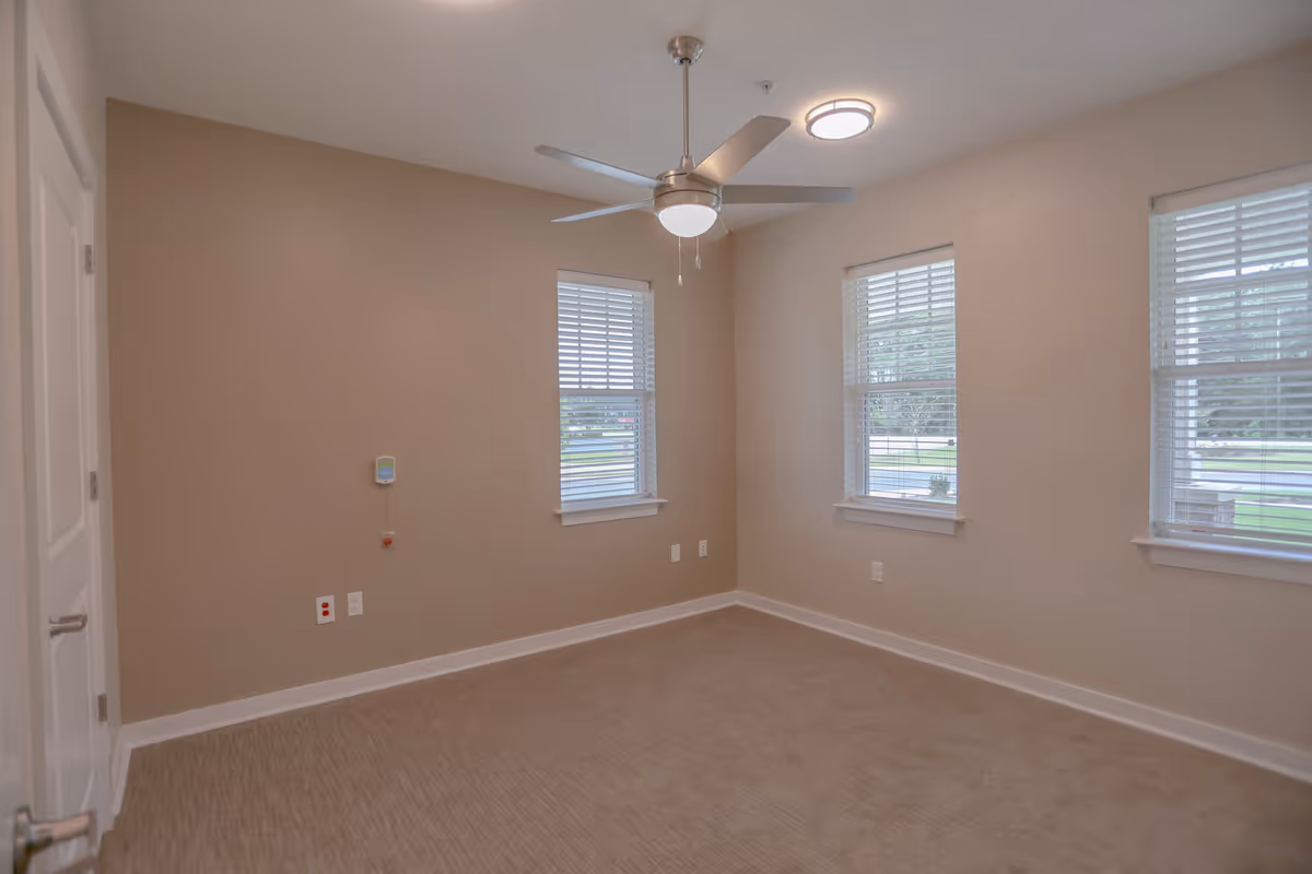 Empty room with beige walls and carpet, three windows with white blinds, a ceiling fan with light, and a closed white door on the left side.