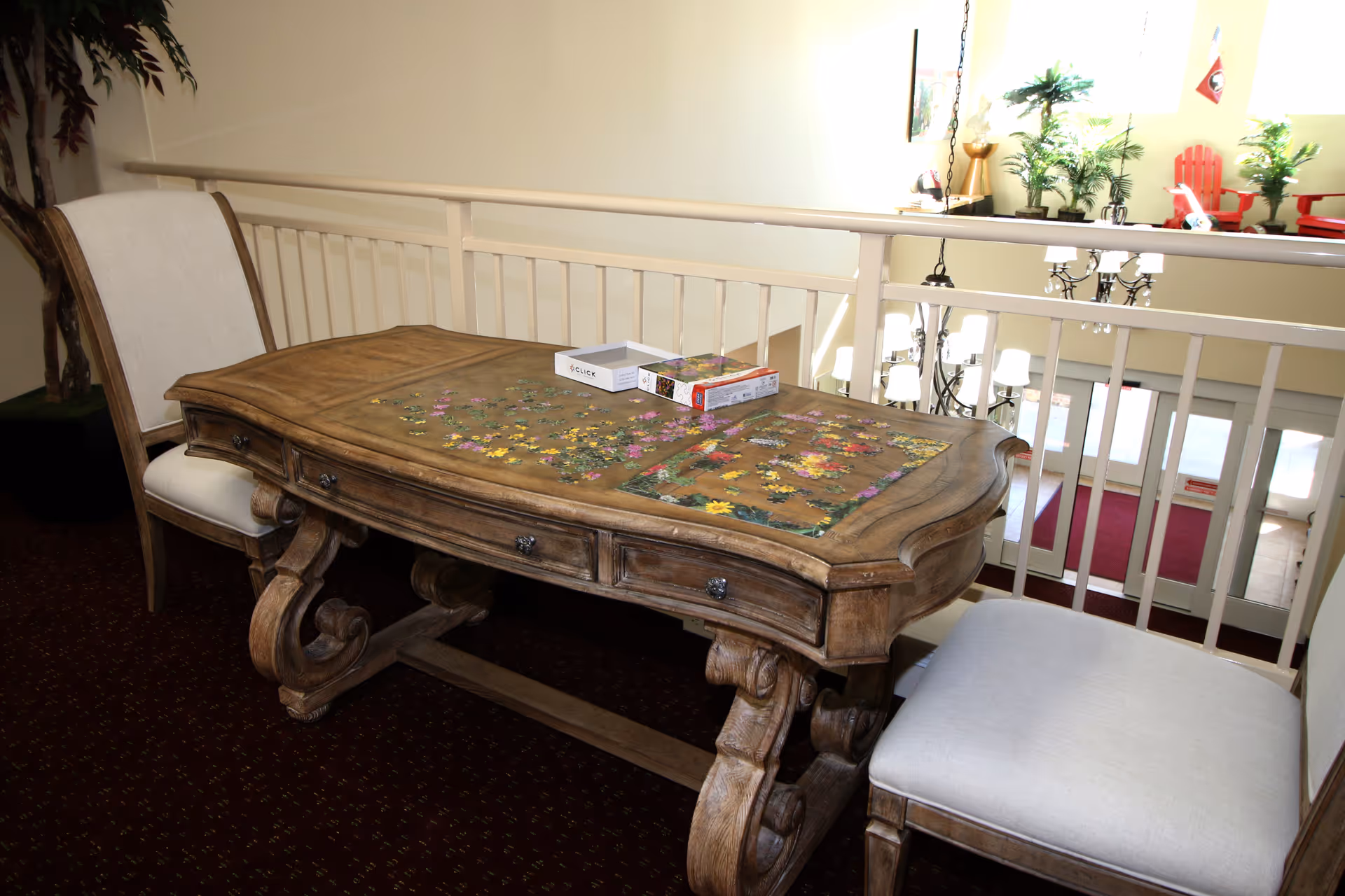 A wooden table with an ornate design and a partially completed floral jigsaw puzzle on top, flanked by two cushioned chairs. The table is positioned near a railing overlooking a lower level with a chandelier and entrance doors visible below. There are plants and red chairs in the background on the upper level.