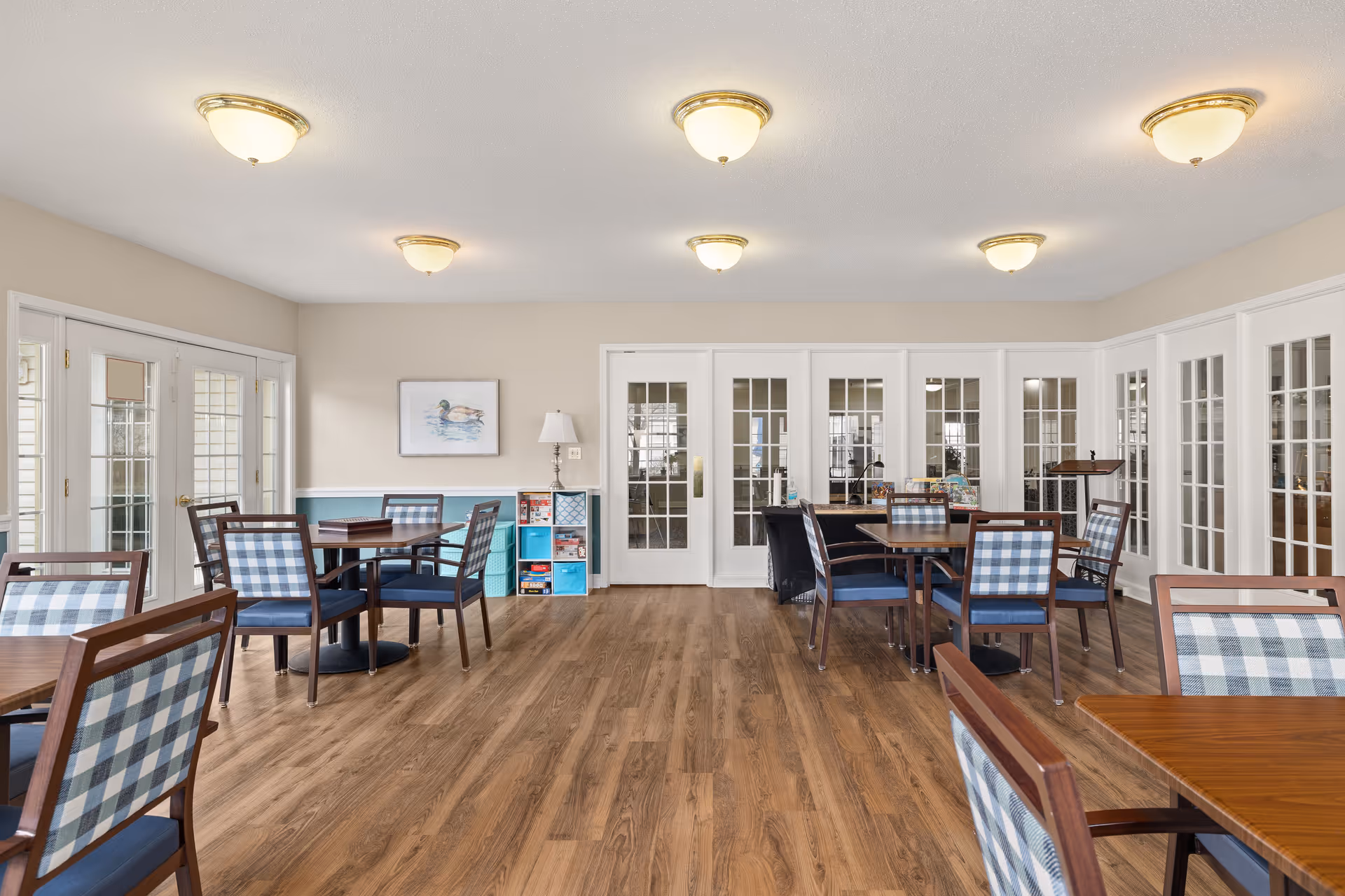 Bright communal dining room with hardwood floors, multiple tables and blue-check upholstered chairs, and glass French doors along the walls.