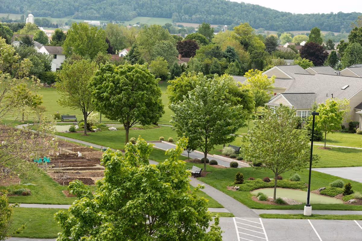 Aerial view of landscaped grounds at Garden Spot Village with walking paths, benches, trees, garden plots, and nearby residential buildings.