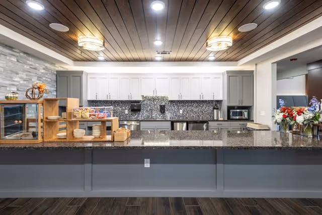 Modern kitchen area with a long granite countertop, gray lower cabinets, white upper cabinets, and a mosaic tile backsplash. The ceiling has wooden panels with recessed lighting and two round ceiling lights. On the countertop, there are wooden display shelves holding plates, bowls, and packaged snacks. To the right, there is a bouquet of flowers in a vase.