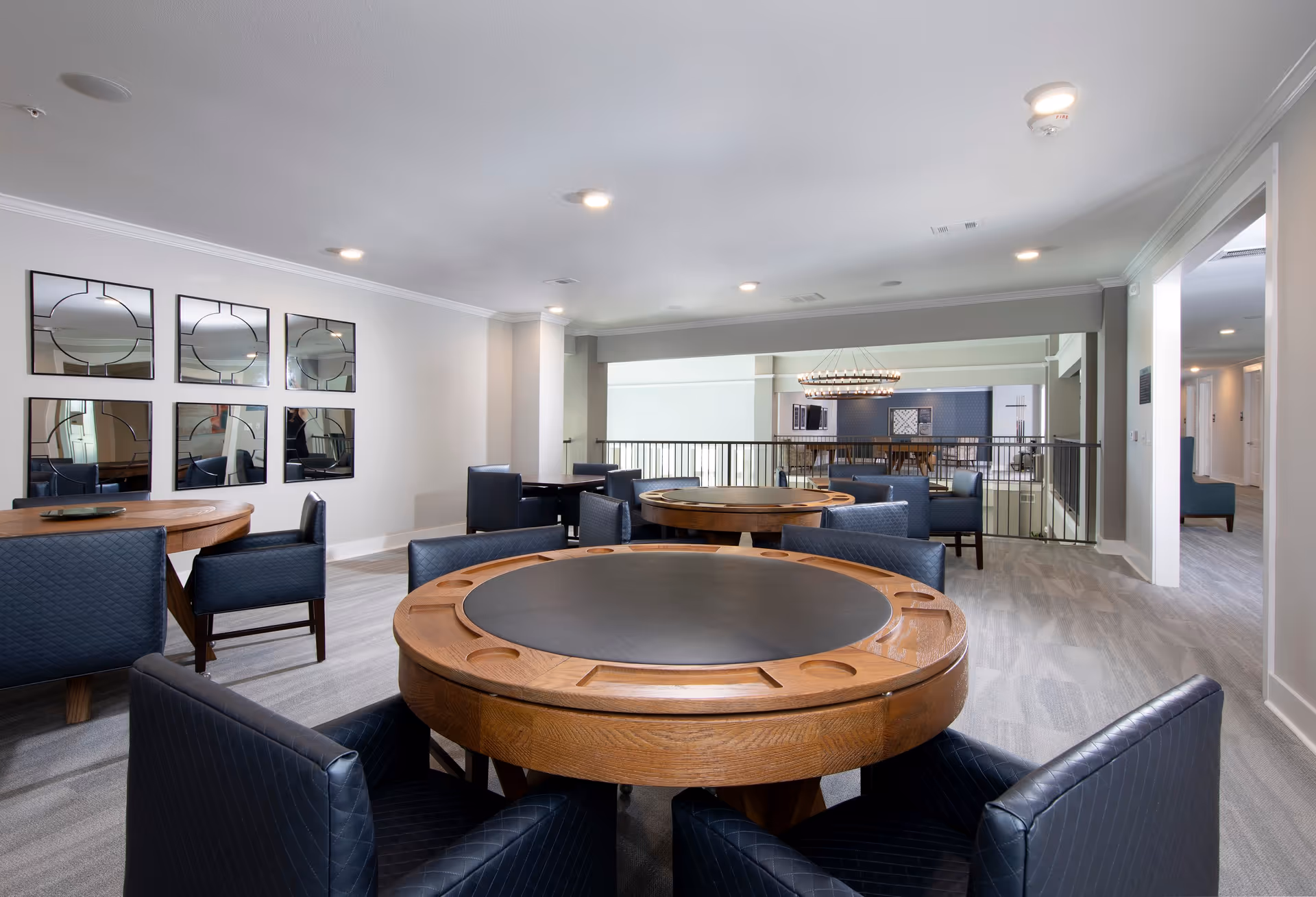 Interior view of a senior living facility common area with round wooden tables featuring black center surfaces and cup holders, surrounded by dark blue cushioned chairs. The room has light gray carpeting, white walls, and decorative mirrors on one wall. In the background, there is a railing overlooking a lower level with additional seating and a large chandelier.