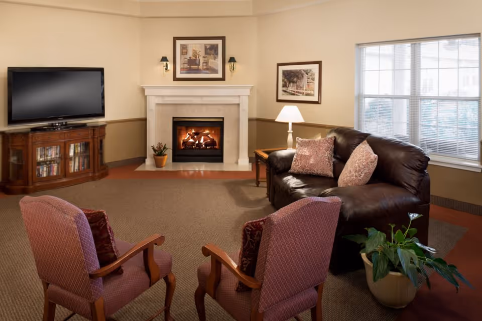 A cozy living room with a lit fireplace in the center, a flat-screen TV on a wooden cabinet to the left, a dark leather sofa with two patterned pillows on the right, two upholstered armchairs facing the fireplace, a side table with a lamp next to the sofa, framed artwork on the walls, a large window with blinds, and a potted plant on the floor.