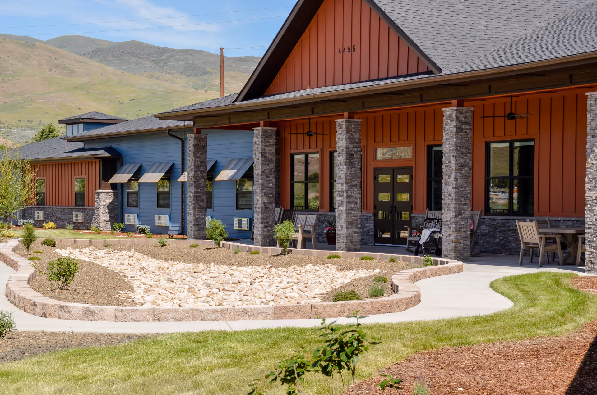 Exterior view of a senior living facility building with a covered porch supported by stone pillars. The building features a combination of orange and blue siding with multiple windows and outdoor seating areas. There is a landscaped area with rocks and small plants in front, and hills are visible in the background under a clear sky.