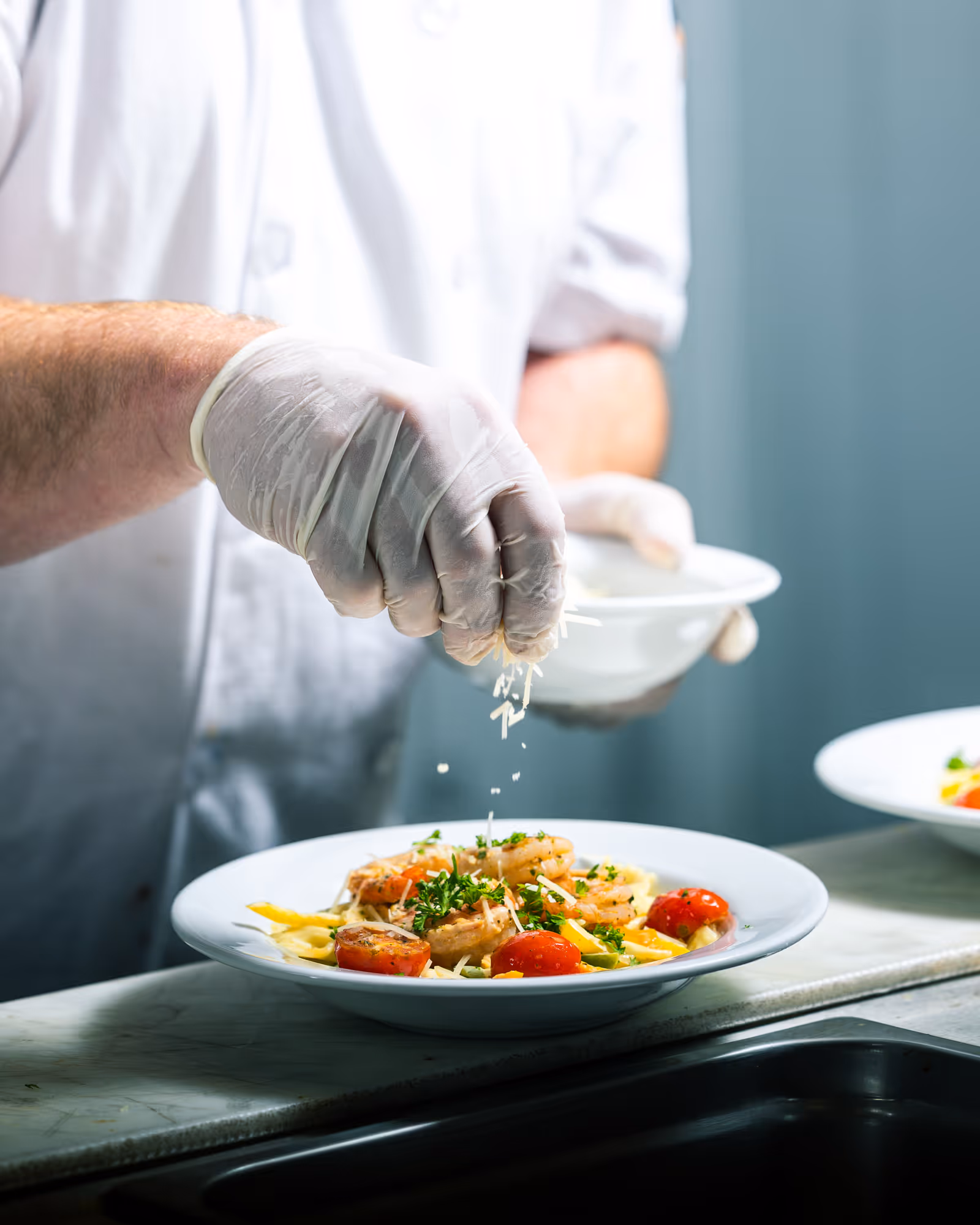 A chef wearing white gloves and a white uniform is sprinkling grated cheese over a plated dish of shrimp, cherry tomatoes, and pasta on a marble countertop.