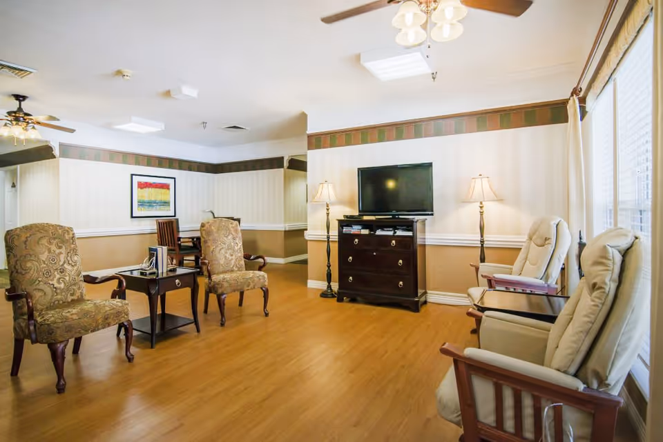 Common living room with upholstered armchairs and recliners arranged around a TV on a dresser, lamps, and large windows.