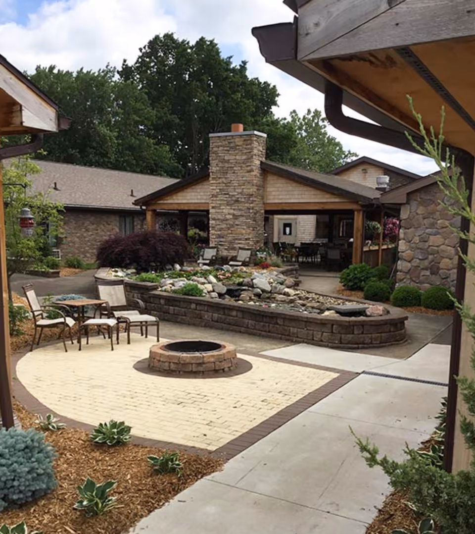 Outdoor courtyard area with a circular brick fire pit surrounded by chairs and a table. There is a raised stone planter with rocks and greenery, and a covered patio with a stone chimney in the background. Trees and shrubs surround the area, creating a peaceful garden setting.