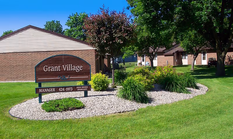 Outdoor view of Grant Village showing a landscaped area with green grass, bushes, and trees. A wooden sign with the text 'Grant Village' and contact information is prominently displayed in front of a brick building under a clear blue sky.