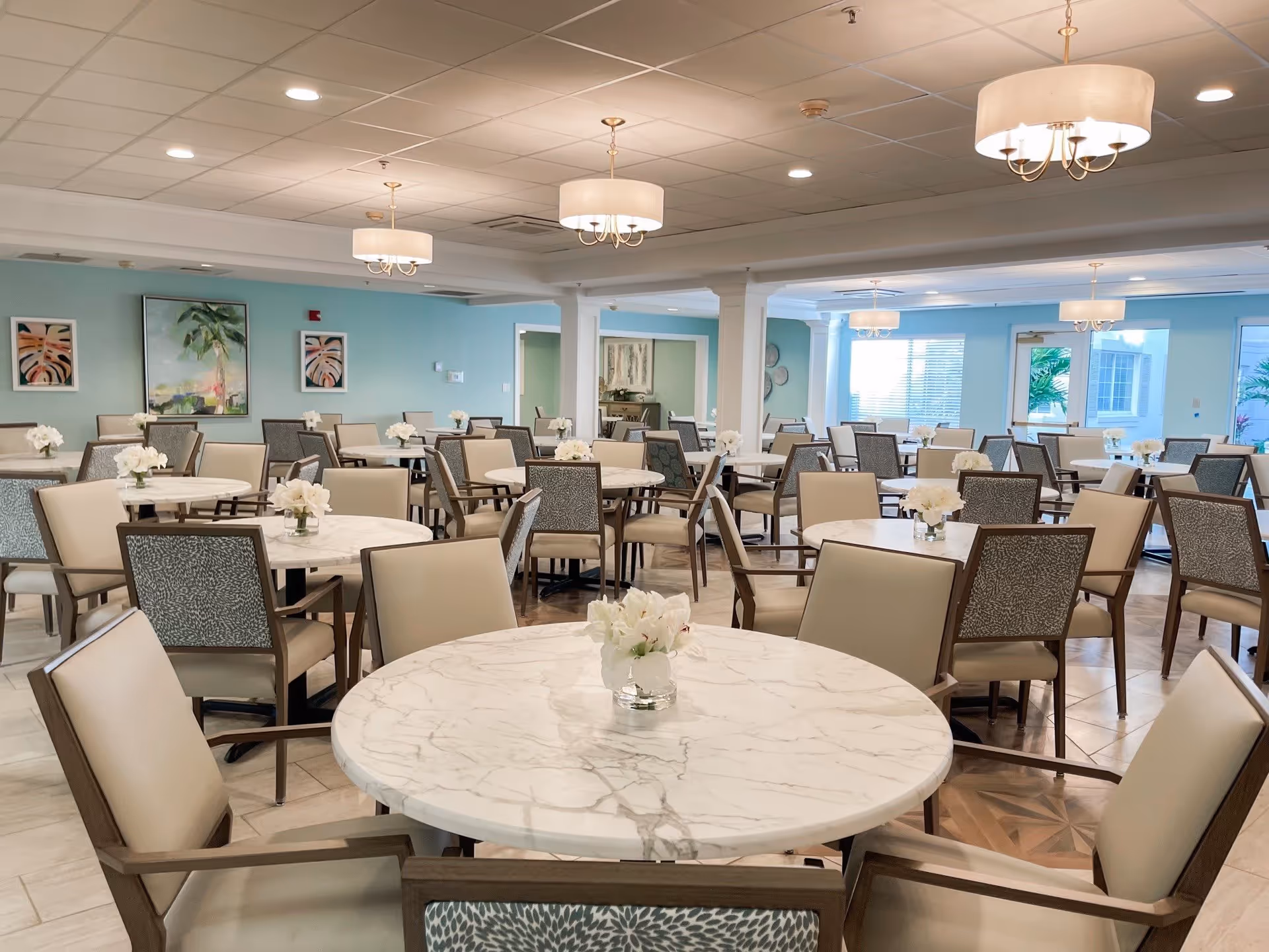 A spacious dining room with multiple round tables topped with white marble surfaces, each adorned with a small vase of white flowers. The room features beige and patterned chairs, light blue walls with framed artwork, and ceiling lights with drum-shaped shades. Large windows allow natural light to brighten the space.