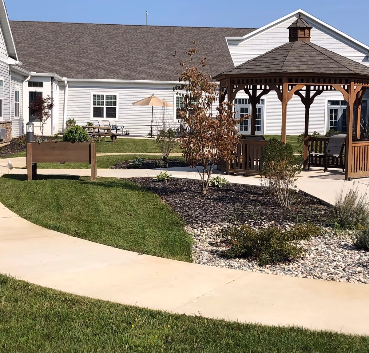 Outdoor garden area at North Woods Village of Kalamazoo featuring a wooden gazebo with chairs, a paved walkway, green grass, small trees, and shrubs. The background shows a white building with multiple windows and doors.