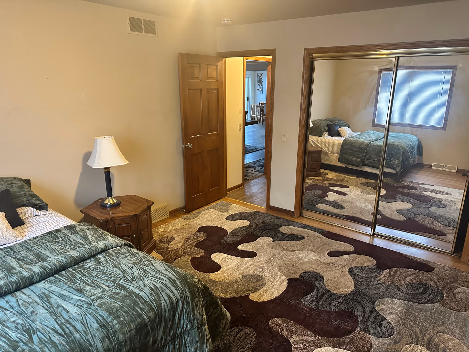 Bedroom with green bedding, a wooden nightstand and lamp, mirrored closet doors, and a large patterned area rug.