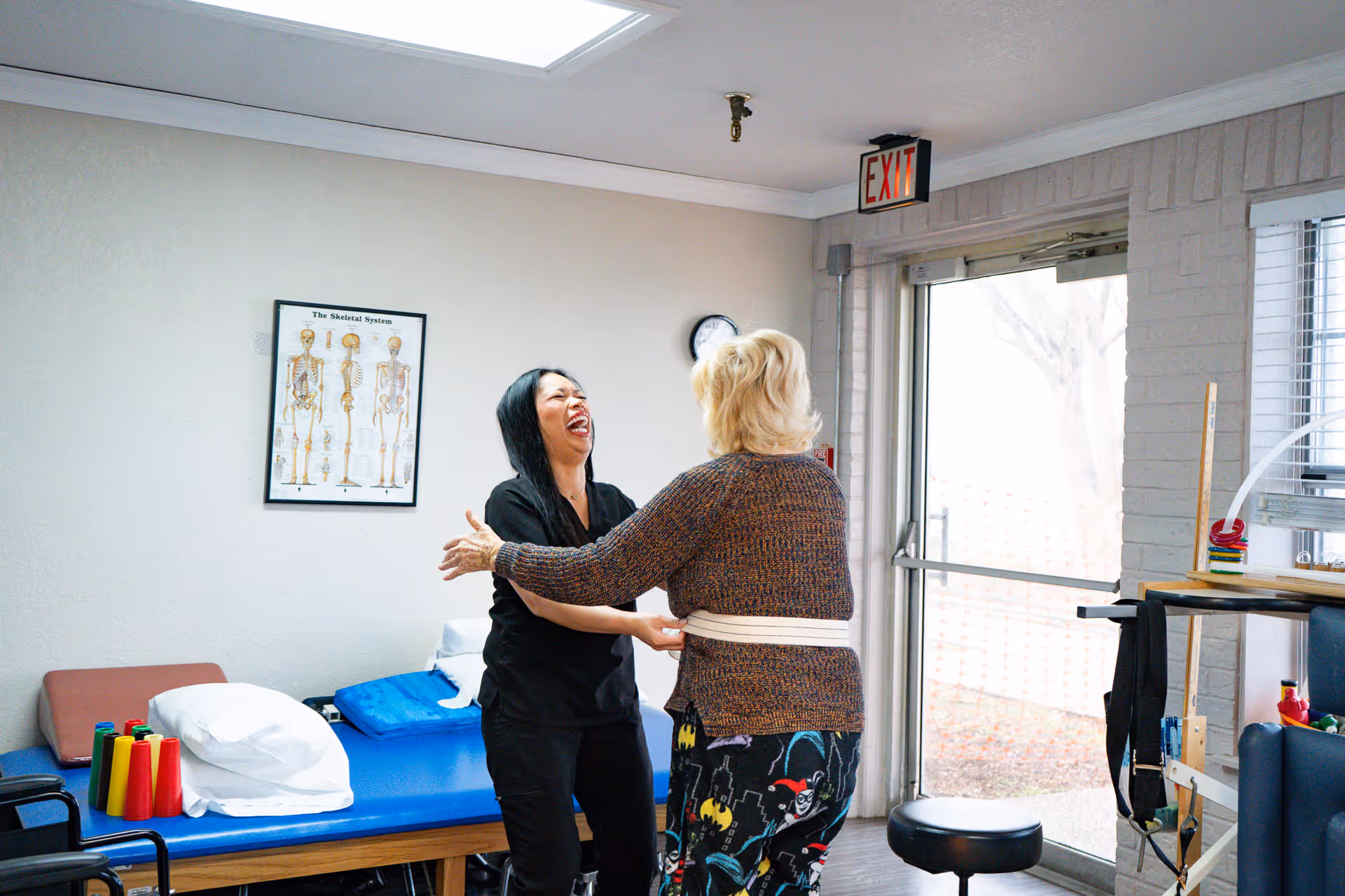 A healthcare worker and an elderly woman are smiling and holding each other in a physical therapy or rehabilitation room. The room has a blue therapy table with pillows, a poster of the skeletal system on the wall, and an exit door with a window letting in natural light.