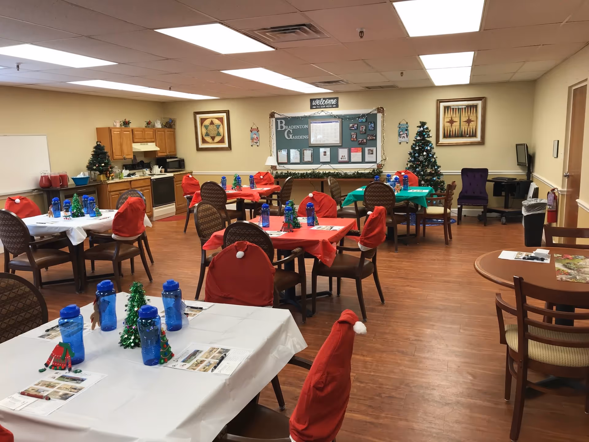 A decorated dining room in a senior living facility with tables covered in red, green, and white tablecloths. Chairs have red Santa hat covers. Blue water bottles and small Christmas tree decorations are on the tables. The room has wooden flooring, a small kitchen area with cabinets, and two decorated Christmas trees. A bulletin board on the wall reads 'Bradenton Gardens' with holiday decorations around it.
