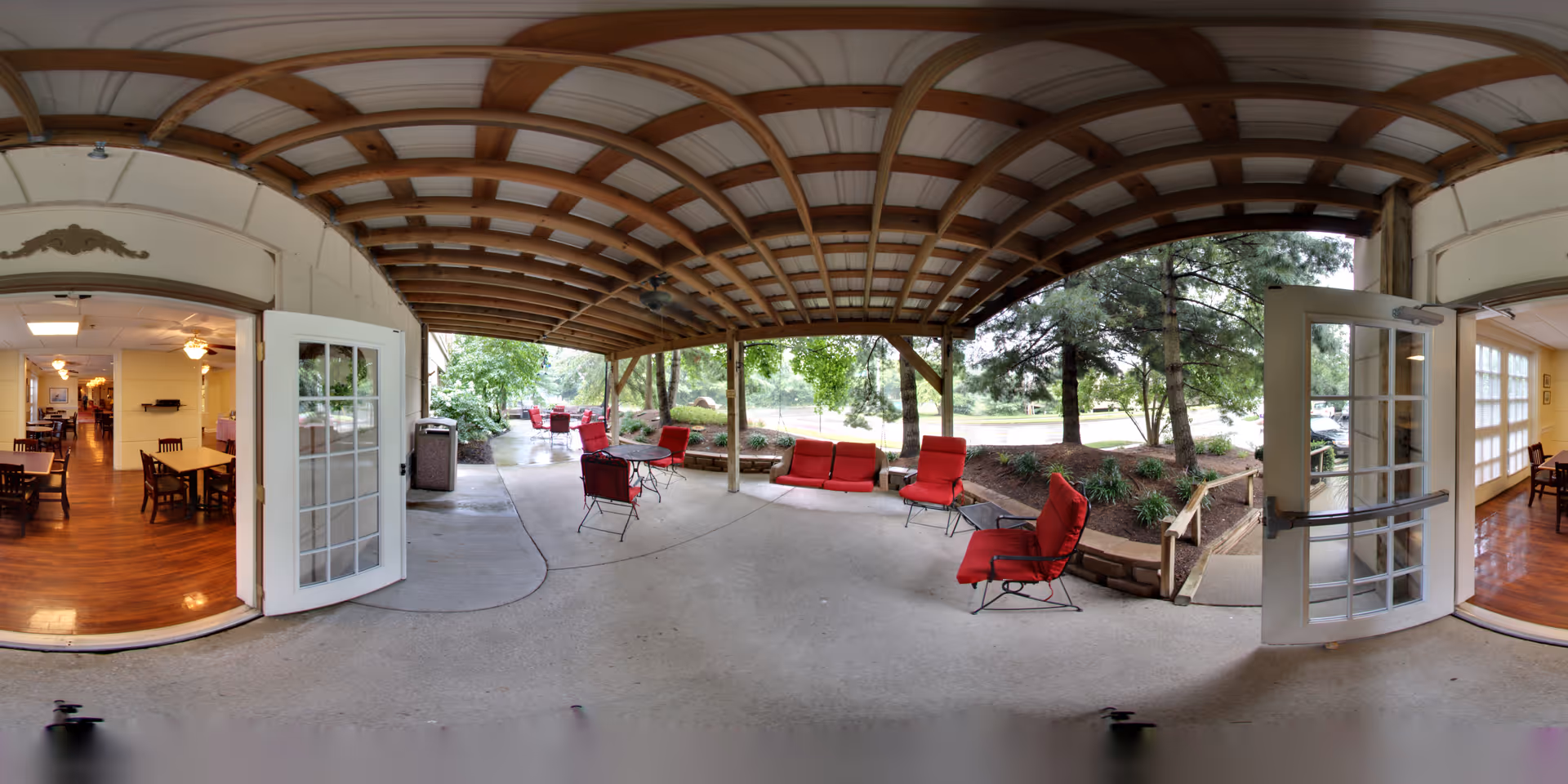 Covered outdoor patio area with red cushioned chairs and tables, surrounded by trees and greenery. Two sets of open double glass doors lead to an indoor dining area with wooden floors and tables with chairs.