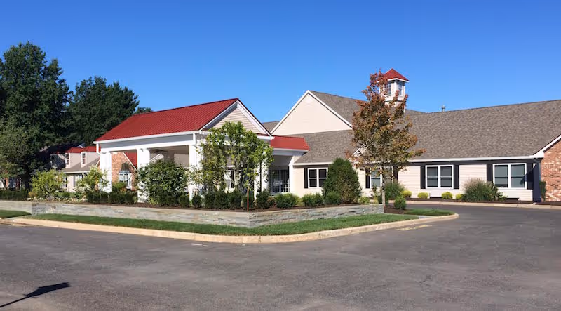 Exterior view of a single-story senior living facility building with a red roofed entrance, beige siding, multiple windows, and landscaped greenery including bushes and trees. The building is surrounded by a paved driveway and parking area under a clear blue sky.
