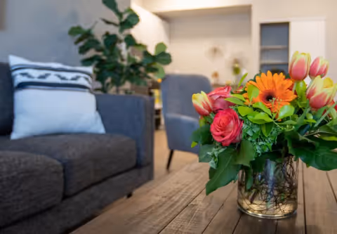 A close-up view of a wooden coffee table with a glass vase holding a colorful bouquet of flowers including orange gerbera daisies, pink roses, and yellow tulips. In the background, there is a gray sofa with a white pillow and a green leafy plant, along with another chair and a softly lit interior space.