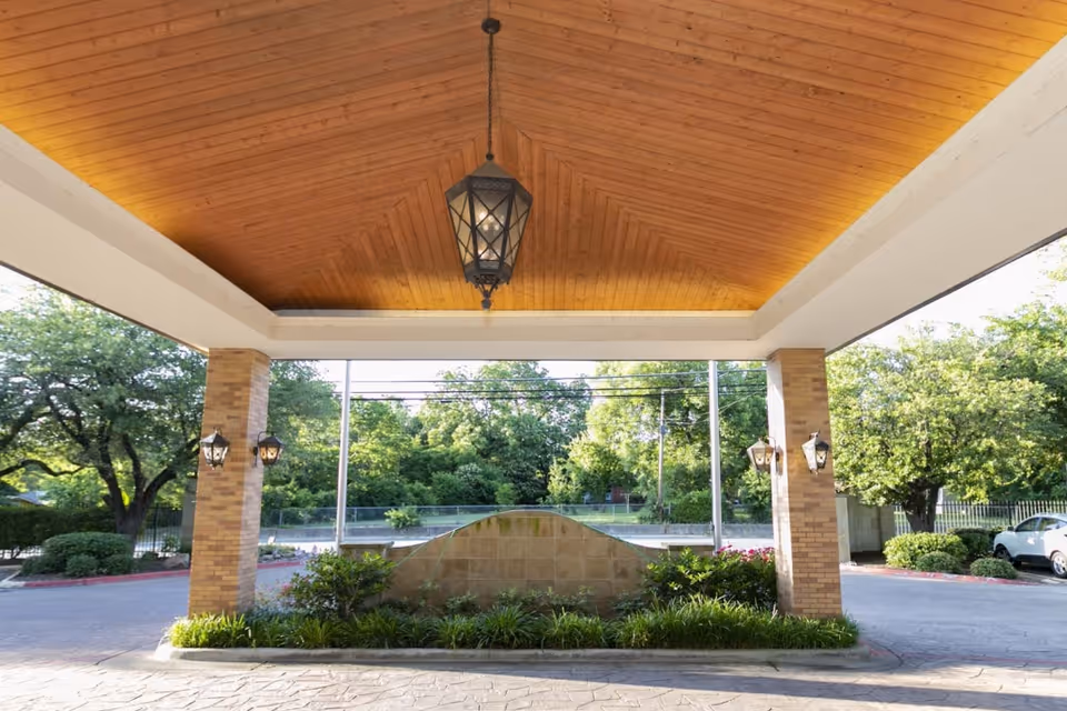 Covered entrance area with a wooden ceiling and hanging lantern light fixture, supported by brick columns. There is landscaping with bushes and plants in front of a low stone wall, and trees and a parked car are visible in the background.